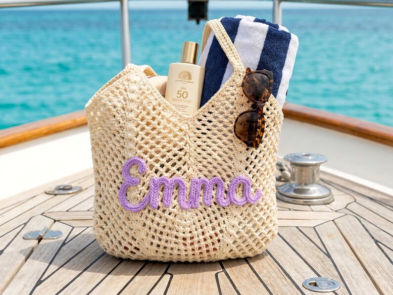 K&ouml;nnte beinhalten: Beige geh&auml;kelte Tragetasche mit dem Namen "Emma" in lila Garn. Die Tasche enth&auml;lt eine Sonnencreme, ein gestreiftes Handtuch und eine Sonnenbrille. Die Tasche befindet sich auf einer Holzoberfl&auml;che mit t&uuml;rkisfarbenem Meer im Hintergrund.
