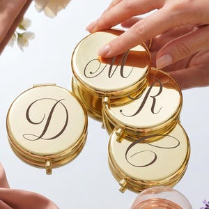 May include: Four round, gold-toned metal compact mirrors with a polished finish. Each mirror features a single, dark, elegant script letter engraved on the lid. The letters visible are D, M, R, and C. The compacts are arranged in a cluster.
