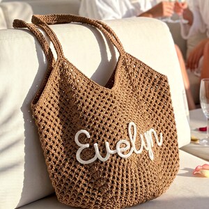 K&ouml;nnte beinhalten: Eine braune geh&auml;kelte Tragetasche mit dem Namen "Evelyn" in wei&szlig;er Schrift gestickt. Die Tasche hat ein gewebtes Muster und zwei Henkel. Die Tasche liegt auf einer wei&szlig;en Oberfl&auml;che.