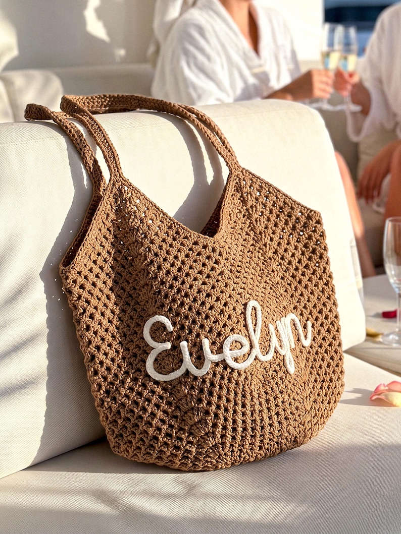K&ouml;nnte beinhalten: Eine braune geh&auml;kelte Tragetasche mit dem Namen "Evelyn" in wei&szlig;er Schrift gestickt. Die Tasche hat ein gewebtes Muster und zwei Henkel. Die Tasche liegt auf einer wei&szlig;en Oberfl&auml;che.