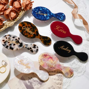 May include: A collection of seven oval hairbrushes in various finishes including blue tortoiseshell, brown tortoiseshell, white tortoiseshell, solid red, solid black, white pearlescent, and pink pearlescent, each personalized with gold script names.