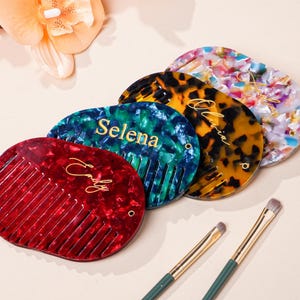 以下が含まれることがあります： 赤、ティール、べっ甲、フローラル柄のパーソナライズされた櫛のコレクション。各櫛には、金色のスクリプトでユニークな名前が付けられています。金色のハンドルと緑色の茎が付いた2本のメイクブラシも存在します。