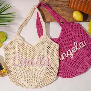 K&ouml;nnte beinhalten: Zwei geh&auml;kelte Tragetaschen, eine cremefarbene und eine pinkfarbene, mit Henkeln. Die cremefarbene Tasche tr&auml;gt den Namen "Camila" in rosa Schrift, und die pinkfarbene Tasche tr&auml;gt den Namen "Angela" in wei&szlig;er Schrift. Eine Flasche Corona Extra Bier und dekorative Fr&uuml;chte sind ebenfalls zu sehen.