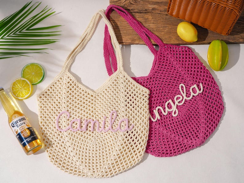 K&ouml;nnte beinhalten: Zwei geh&auml;kelte Tragetaschen, eine cremefarbene und eine pinkfarbene, mit Henkeln. Die cremefarbene Tasche tr&auml;gt den Namen "Camila" in rosa Schrift, und die pinkfarbene Tasche tr&auml;gt den Namen "Angela" in wei&szlig;er Schrift. Eine Flasche Corona Extra Bier und dekorative Fr&uuml;chte sind ebenfalls zu sehen.