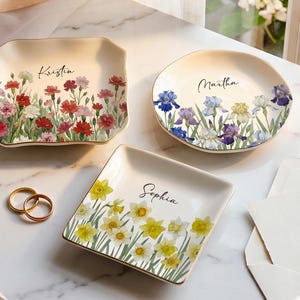 Könnte beinhalten: Drei Keramikschalen mit goldfarbenen Rändern. Eine quadratische Schale zeigt rote und rosa Nelken sowie den Namen Kristin. Eine runde Schale zeigt blaue und violette Schwertlilien sowie den Namen Martha. Eine quadratische Schale zeigt gelbe Narzissen und den Namen Sophia.