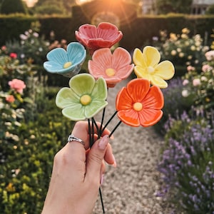 Könnte beinhalten: Ein Strauß aus sechs Keramikblumen an dünnen grünen Metallstielen. Die Blumen haben glänzende Blütenblätter in den Farben Rot, Hellblau, Pfirsich, Hellgrün, Gelb und Orange, jede mit einer kleinen gelben Mitte, in einem Bündel angeordnet.