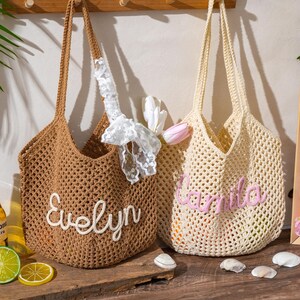 K&ouml;nnte beinhalten: Zwei geh&auml;kelte Tragetaschen. Eine ist braun mit dem Namen "Evelyn" in wei&szlig;er Schrift. Die andere ist cremefarben mit dem Namen "Camila" in rosa Schrift. Beide Taschen haben lange Henkel und ein gewebtes Design. Dekorative Elemente sind um die Taschen platziert.