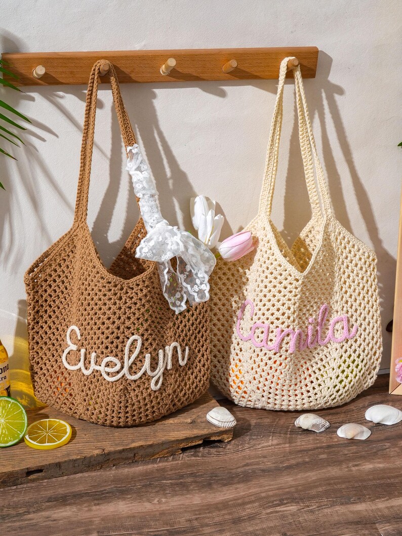 K&ouml;nnte beinhalten: Zwei geh&auml;kelte Tragetaschen. Eine ist braun mit dem Namen "Evelyn" in wei&szlig;er Schrift. Die andere ist cremefarben mit dem Namen "Camila" in rosa Schrift. Beide Taschen haben lange Henkel und ein gewebtes Design. Dekorative Elemente sind um die Taschen platziert.