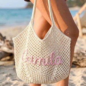 K&ouml;nnte beinhalten: Eine cremefarbene, offen gewebte H&auml;keltasche mit einem rosa gestickten "Camila" auf der Vorderseite. Die Tasche hat zwei cremefarbene Griffe und wird an einem Strand gehalten. Die Tasche ist ein Strandaccessoire.