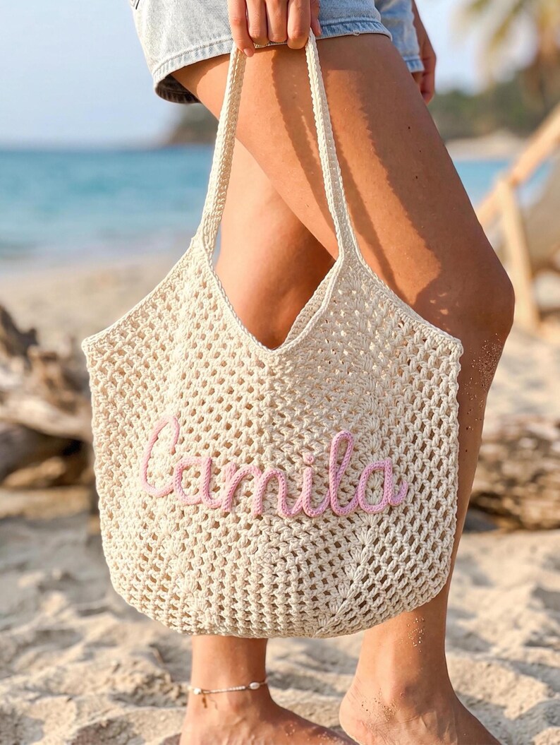 K&ouml;nnte beinhalten: Eine cremefarbene, offen gewebte H&auml;keltasche mit einem rosa gestickten "Camila" auf der Vorderseite. Die Tasche hat zwei cremefarbene Griffe und wird an einem Strand gehalten. Die Tasche ist ein Strandaccessoire.