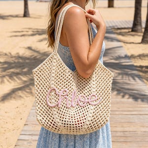 K&ouml;nnte beinhalten: Eine beigefarbene, offen gewebte Tragetasche mit einem rosa gestickten "Chloe"-Schriftzug. Die Tasche hat zwei Schultergurte und wird von einer Person am Strand gehalten. Die Tasche ist hell und scheint aus einem gewebten Material zu sein.