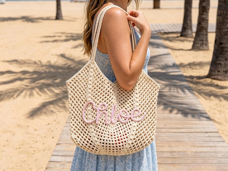 K&ouml;nnte beinhalten: Eine beigefarbene, offen gewebte Tragetasche mit einem rosa gestickten "Chloe"-Schriftzug. Die Tasche hat zwei Schultergurte und wird von einer Person am Strand gehalten. Die Tasche ist hell und scheint aus einem gewebten Material zu sein.