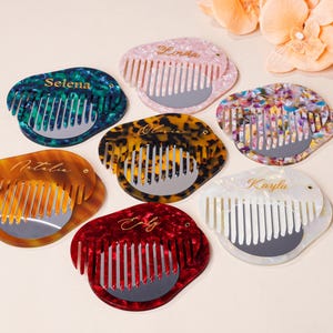 May include: A selection of personalized combs in shades like green, pink, and red. Each comb has a mirror and a name in script. The combs display a tortoiseshell pattern and are arranged on a light surface.