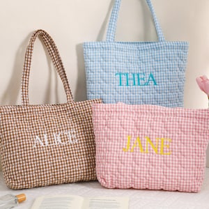 May include: Three quilted tote bags in brown, blue, and pink gingham patterns. Each bag has a different name embroidered on it: "ALICE", "THEA", and "JANE". The bags have matching handles and are displayed on a white surface.