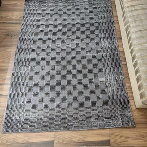 May include: A rectangular gray rug with a checkered pattern. The rug is made of a soft material and has a textured appearance. The rug is placed on a wooden floor.