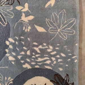 Könnte beinhalten: Ein blauer Teppich mit floralem Design. Der Teppich hat weiße und braune Blumen- und Blättermuster auf hellblauem Hintergrund. Das Design umfasst stilisierte Bäume und abstrakte Formen.