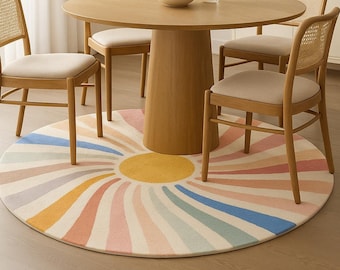 Op de afbeelding: Een rond tapijt met een zonnestraalpatroon in pastelkleuren, waaronder roze, geel, blauw en oranje. Een houten eettafel en vier stoelen met geweven rugleuningen en beige kussens zijn erboven geplaatst.