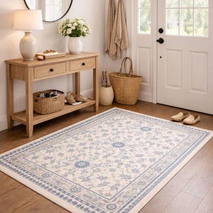 Peut inclure: Tapis rectangulaire à motifs floraux bleus et beiges. Il est posé sur un parquet dans une entrée lumineuse, avec une console en bois, un miroir et une porte en arrière-plan. Dimensions approximatives : 180 x 270 cm.