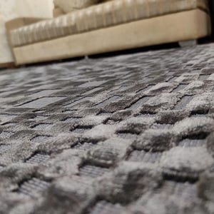 May include: A close-up shot of a gray area rug with a textured, geometric pattern. The rug's design features a series of raised, rounded shapes, creating a tactile surface. The color palette is a blend of gray tones, adding depth and dimension to the rug.