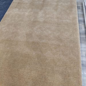 May include: A large, rectangular, tan-colored rug with a soft, plush texture. The rug is displayed on a light-colored wooden floor, showcasing its neutral color and simple design. The rug's dimensions are not visible.