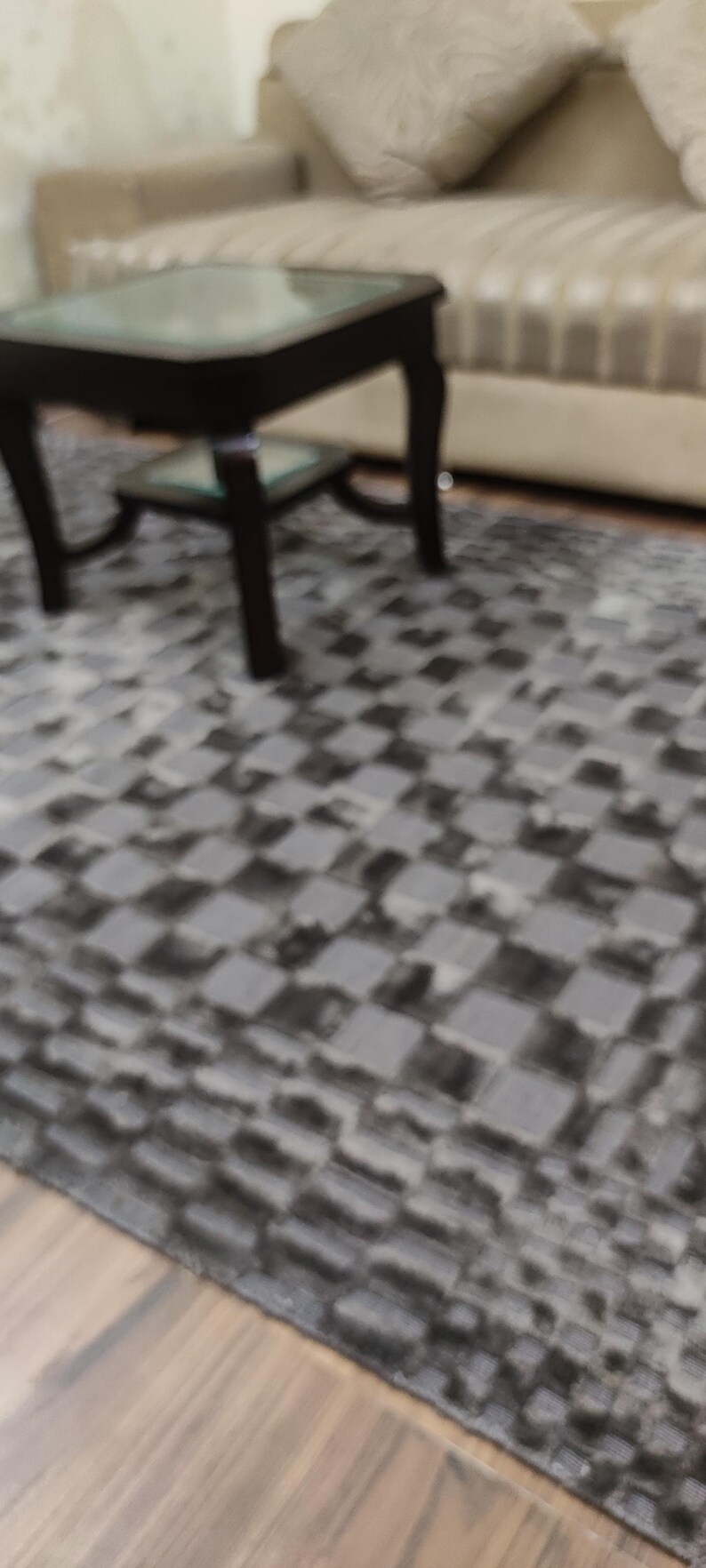 May include: A living room scene featuring a dark wood coffee table with a glass top, a beige sofa with pillows, and a gray checkered rug. The rug covers the floor in front of the sofa and table.