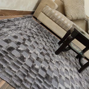 May include: A rectangular area rug with a geometric pattern in shades of gray and black. The rug has a textured appearance and is placed on a wooden floor. A dark brown coffee table and a beige sofa are in the background.