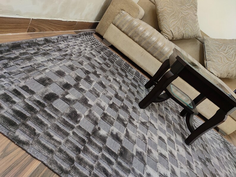 May include: A rectangular area rug with a geometric pattern in shades of gray and black. The rug has a textured appearance and is placed on a wooden floor. A dark brown coffee table and a beige sofa are in the background.
