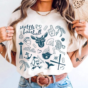 May include: Ivory-colored t-shirt with a navy blue graphic design featuring Seattle baseball-themed illustrations. The design includes text that reads "Seattle Baseball" along with images of a moose, baseballs, and other related icons.