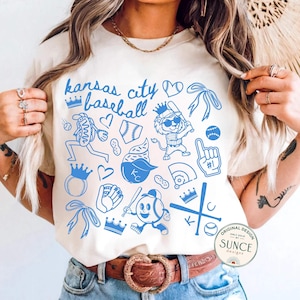 model wearing ivory comfort colors shirt that says kansas city baseball and has a baseball doodle design