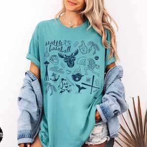 May include: Seafoam green t-shirt with a navy blue graphic design featuring Seattle landmarks and baseball-themed illustrations. The text "Seattle Baseball" is at the top. The shirt is a comfortable, casual style.