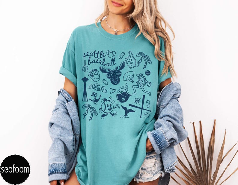 May include: Seafoam green t-shirt with a navy blue graphic design featuring Seattle landmarks and baseball-themed illustrations. The text "Seattle Baseball" is at the top. The shirt is a comfortable, casual style.