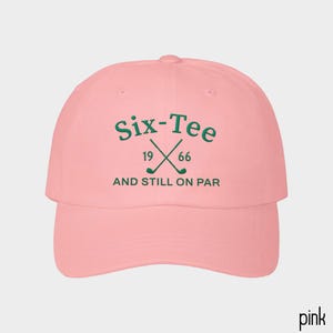 May include: A light pink baseball cap featuring dark green embroidered text that reads Six-Tee, 19 66, and AND STILL ON PAR, with a graphic of two crossed golf clubs positioned between the numbers 19 and 66 on the front panel.
