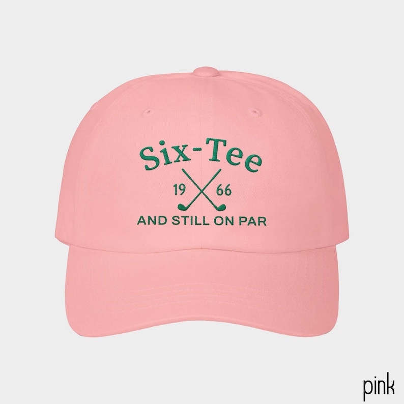 May include: A light pink baseball cap featuring dark green embroidered text that reads Six-Tee, 19 66, and AND STILL ON PAR, with a graphic of two crossed golf clubs positioned between the numbers 19 and 66 on the front panel.