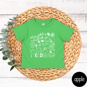 May include: A bright green t-shirt with white graphic illustrations and the words "philadelphia football". The design includes footballs, a helmet, an eagle, and other sports-related images. The shirt is displayed on a woven, circular mat.