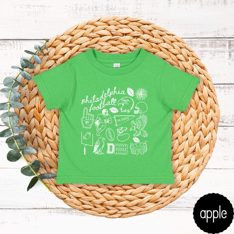 May include: A bright green t-shirt with white graphic illustrations and the words "philadelphia football". The design includes footballs, a helmet, an eagle, and other sports-related images. The shirt is displayed on a woven, circular mat.