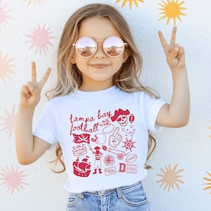 May include: A white short-sleeved t-shirt featuring a red graphic print with the text "tampa bay football" and various illustrations including a pirate skull, a ship, a football helmet, a cannon, a foam finger, and a stadium.