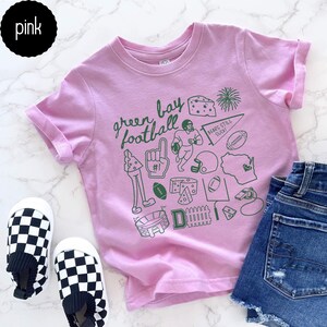 May include: A pale pink t-shirt featuring the text "green bay football" and green illustrations of football-related items and Wisconsin. The shirt is shown with denim shorts and chequered shoes.