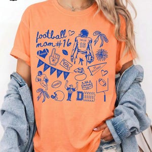 May include: An orange short-sleeved t-shirt featuring a blue graphic print with the text football mom #16, Taylor 16, and Go Bulldogs! The design includes various football-themed illustrations like a player, whistle, pennant, and goal post.