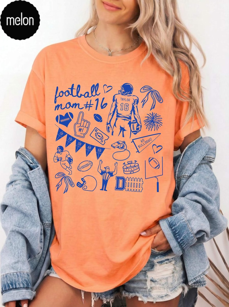 May include: An orange short-sleeved t-shirt featuring a blue graphic print with the text football mom #16, Taylor 16, and Go Bulldogs! The design includes various football-themed illustrations like a player, whistle, pennant, and goal post.