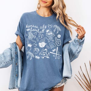 May include: Light blue t-shirt featuring the text "Kansas City Baseball" and white baseball-themed illustrations. The design includes baseballs, crowns, and a baseball player. The shirt is a light blue colour.
