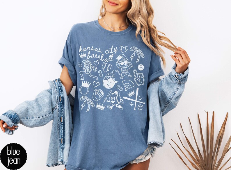 May include: Light blue t-shirt featuring the text "Kansas City Baseball" and white baseball-themed illustrations. The design includes baseballs, crowns, and a baseball player. The shirt is a light blue colour.