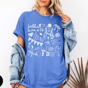 May include: A blue t-shirt featuring a white graphic print with the text football mom #16, surrounded by various football-themed illustrations including a player, a helmet, a foam finger, a whistle, a pennant, and a stadium goal post.