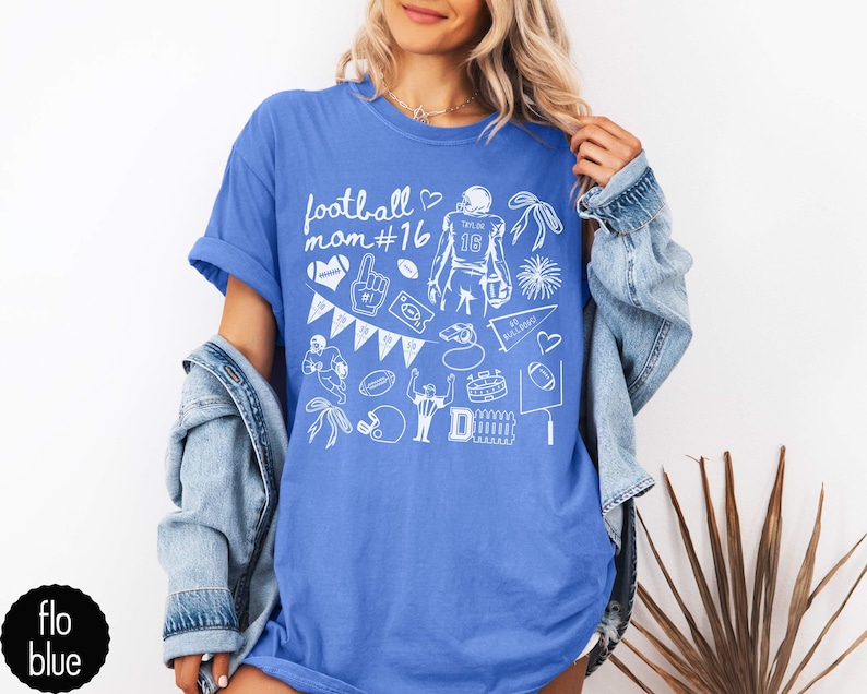 May include: A blue t-shirt featuring a white graphic print with the text football mom #16, surrounded by various football-themed illustrations including a player, a helmet, a foam finger, a whistle, a pennant, and a stadium goal post.