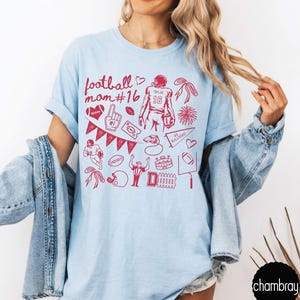May include: A light blue t-shirt featuring a red graphic print with the text football mom #16, a football player, a pennant, a whistle, a stadium, a referee, a helmet, hearts, bows, and various football-themed icons arranged in a collage pattern.