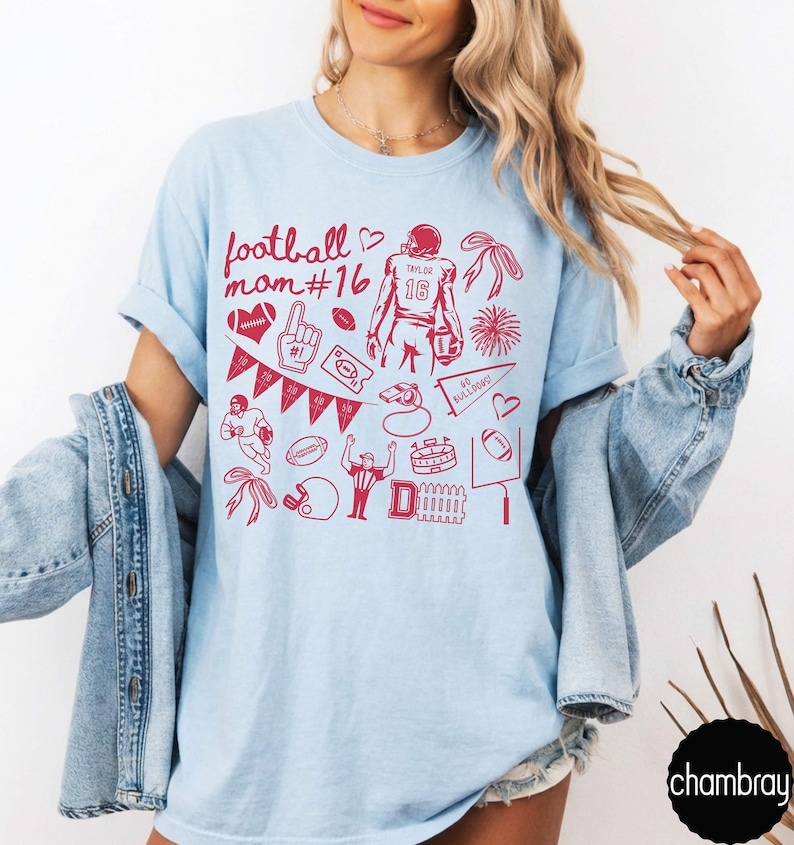May include: A light blue t-shirt featuring a red graphic print with the text football mom #16, a football player, a pennant, a whistle, a stadium, a referee, a helmet, hearts, bows, and various football-themed icons arranged in a collage pattern.