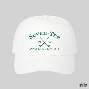 May include: A white baseball cap featuring green embroidered text that reads Seven-Tee, the numbers 19 and 56, and the phrase AND STILL ON PAR. The design includes a graphic of two crossed golf clubs centered between the numbers.