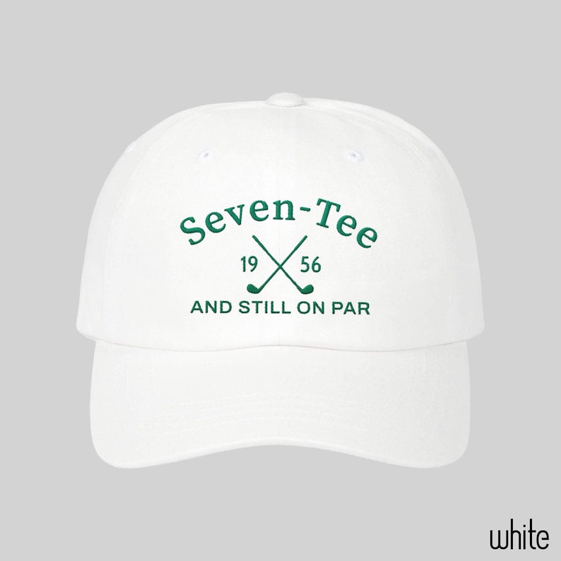 May include: A white baseball cap featuring green embroidered text that reads Seven-Tee, the numbers 19 and 56, and the phrase AND STILL ON PAR. The design includes a graphic of two crossed golf clubs centered between the numbers.