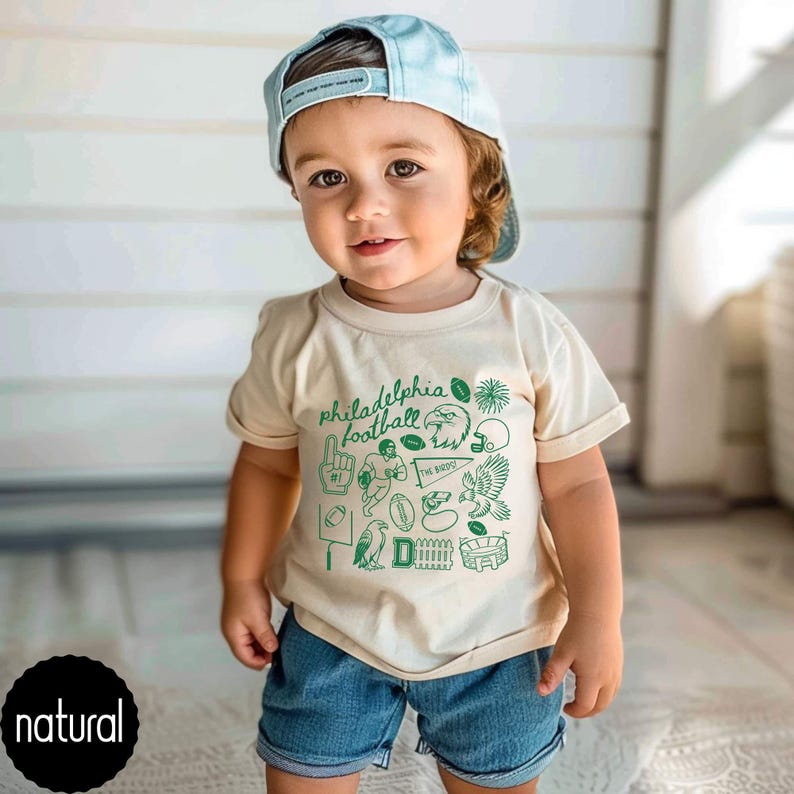 May include: A toddler wearing a light beige t-shirt with a green graphic design that says "Philadelphia football". The design includes footballs, an eagle, and a stadium. The child is also wearing blue denim shorts and a light blue baseball cap.