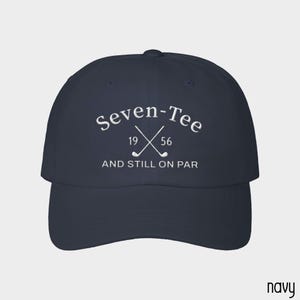 May include: A navy blue baseball cap featuring white embroidered text that reads Seven-Tee, the numbers 19 and 56, and the phrase AND STILL ON PAR, accompanied by a graphic of two crossed golf clubs centered on the front panel of the hat.