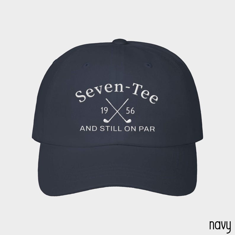 May include: A navy blue baseball cap featuring white embroidered text that reads Seven-Tee, the numbers 19 and 56, and the phrase AND STILL ON PAR, accompanied by a graphic of two crossed golf clubs centered on the front panel of the hat.